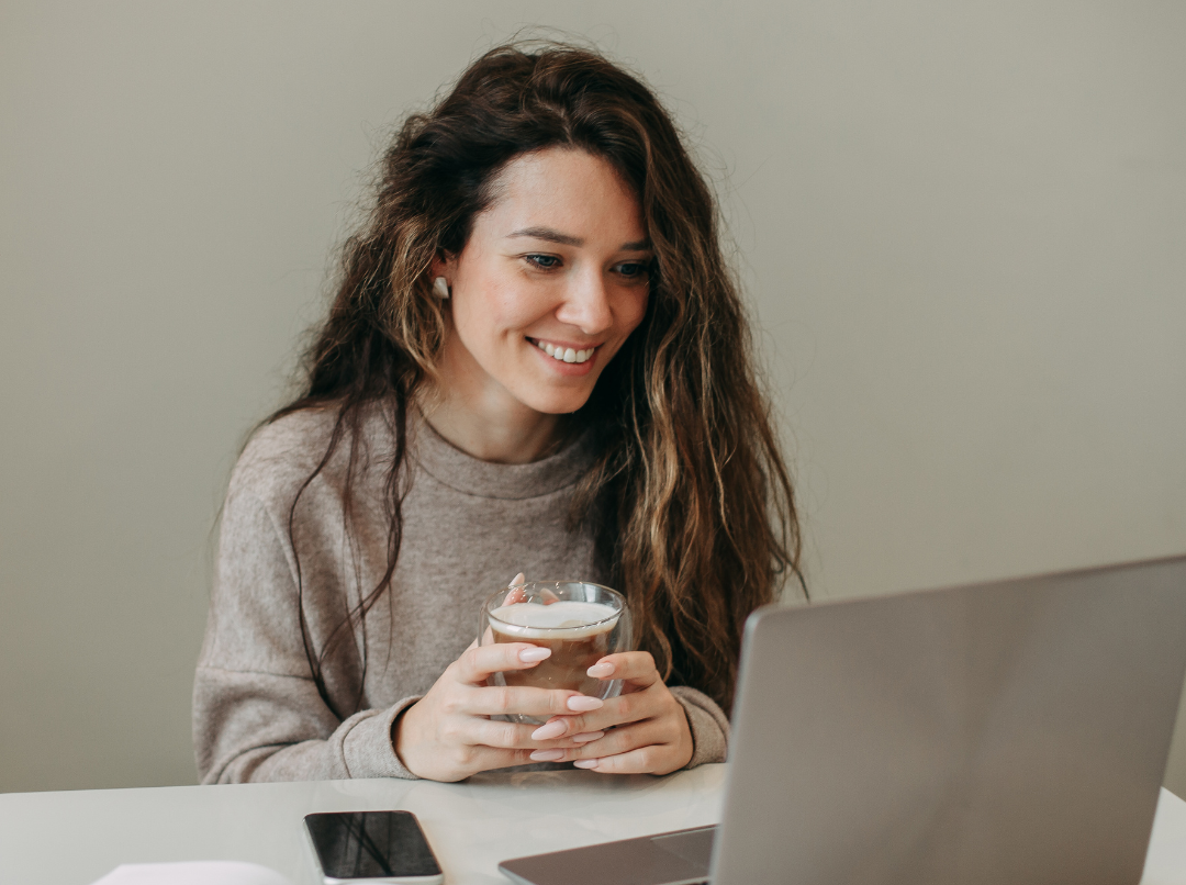 A smiling PhD student looking at a laptop, holding a coffee cup.