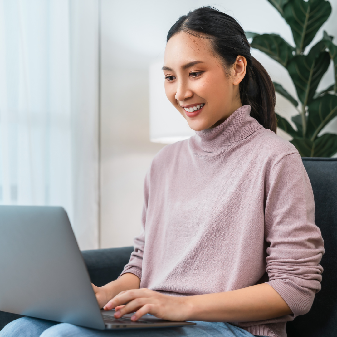 A seated woman smiles while looking at and typing on her laptop in a modern lobby.