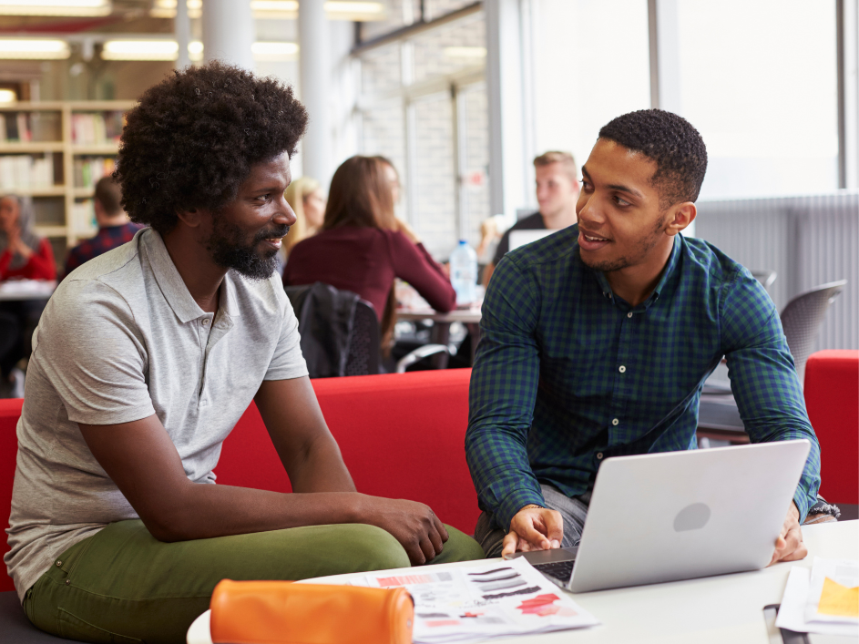 Two students on a couch, conversing while reviewing a laptop and report paper.