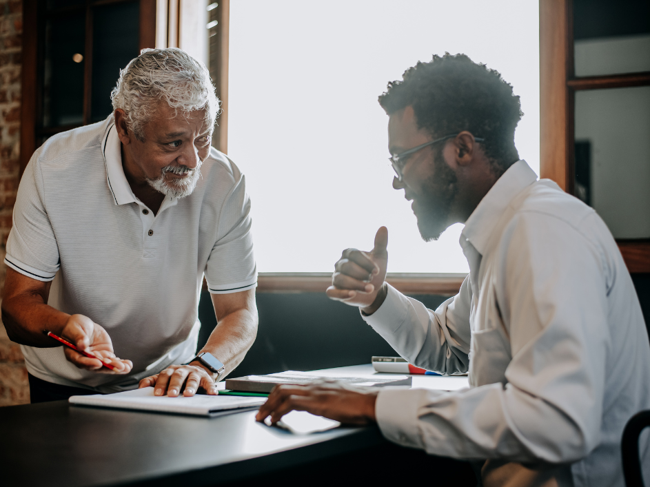 An older advisor speaking to a PhD student sitting at a desk with documents.