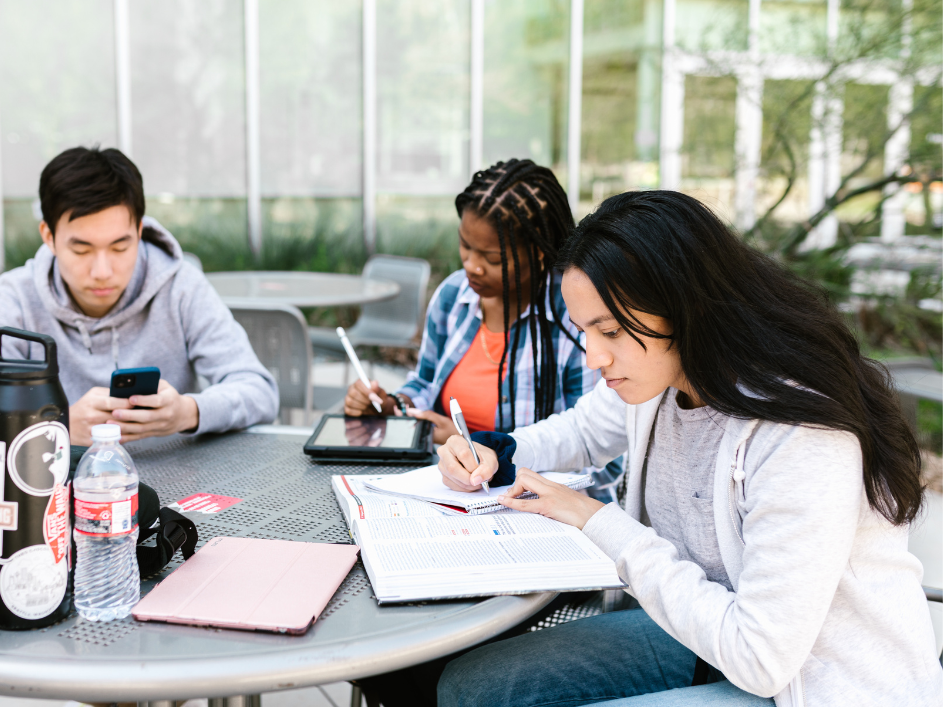 Three PhD students writing notes at an outdoor table.