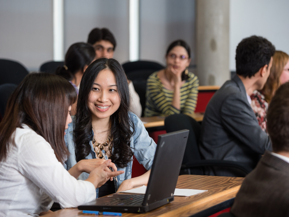 Two PhD students discussing at shared desk with a laptop.