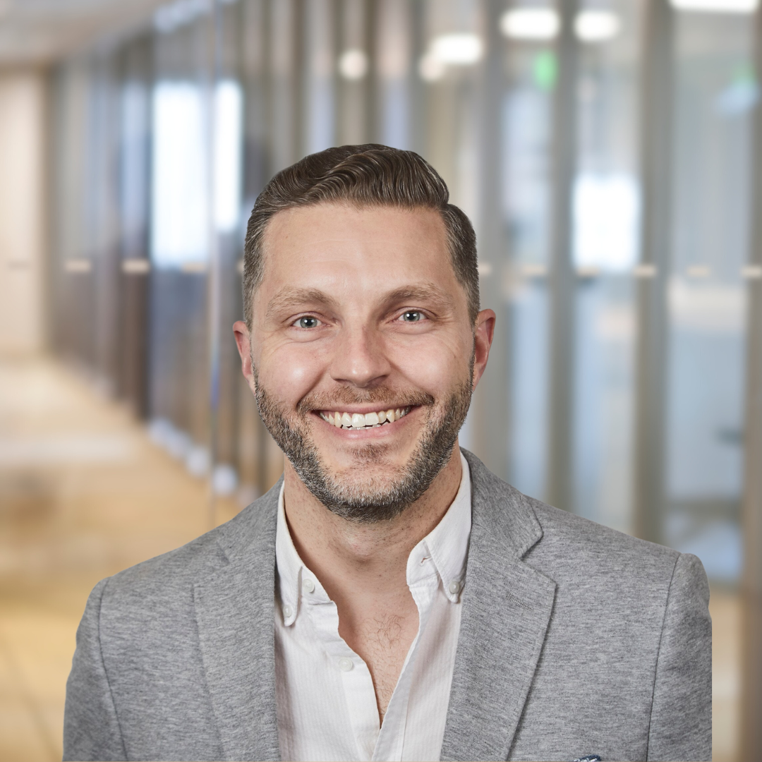 Headshot of Shane Saunderson, a smiling man with a beard, short, brown hair, wearing a grey suit.