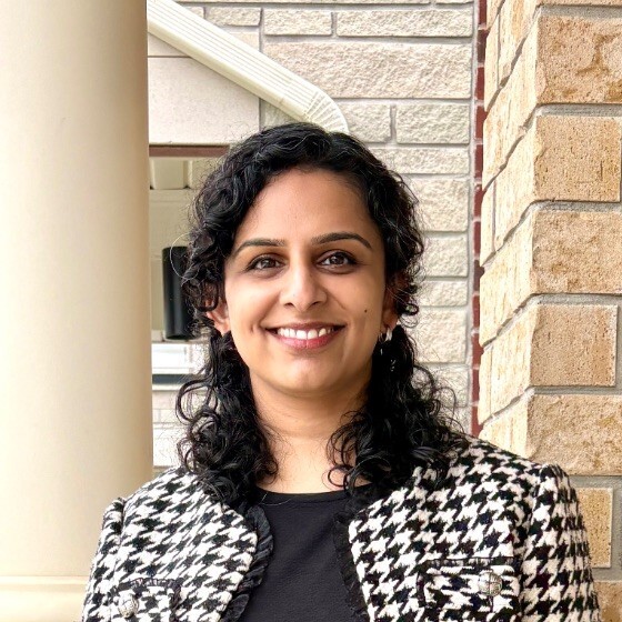 Headshot of Pavithra Balaji, a smiling woman with dark hair, wearing a checkered coat over a black shirt.