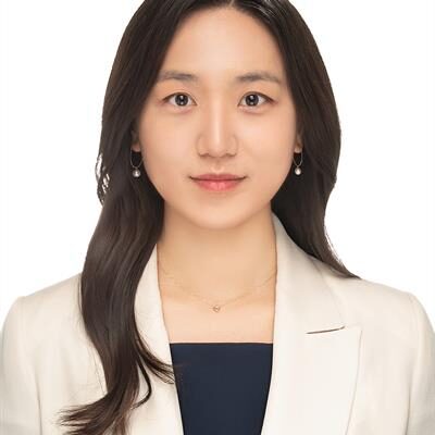 A headshot of Judy Han, a smiling woman with dark hair, wearing a white blazer over a black shirt.