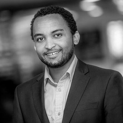 A black-and-white headshot of Addisu Lashitea, a smiling man with dark hair, wearing a blazer and a collared shirt.